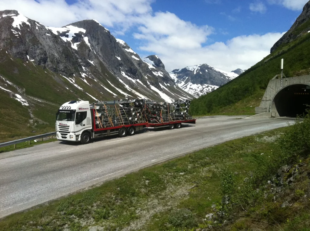 BK Hengerens lastebil langs veien i fjellheimen, blå himmel med skyer og snødekte fjell i bakkant.